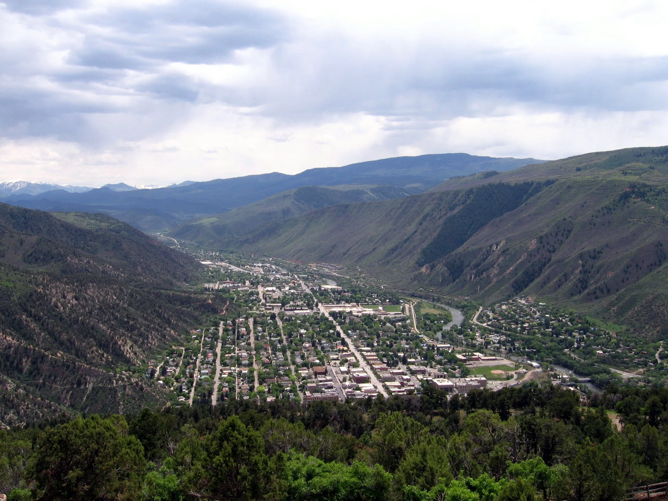 A small town situated in a valley surrounded by green mountains under a cloudy sky.