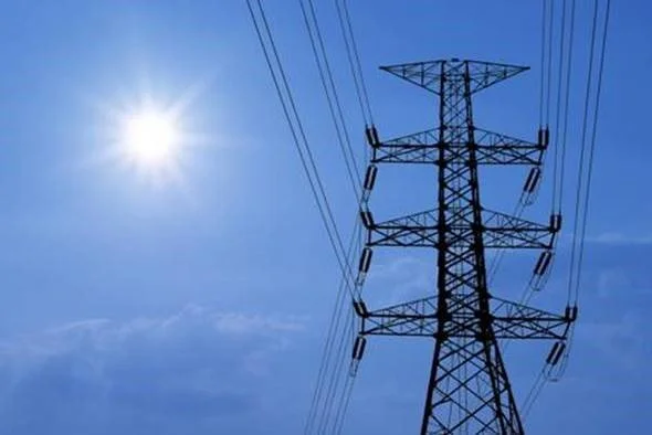 A tall electricity pylon with power lines against a bright sun and blue sky.