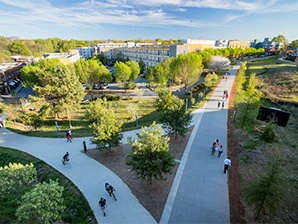 A park with winding walking paths, green trees, and people walking and biking, with residential buildings in the background on a sunny day.