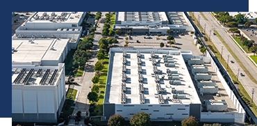 Aerial view of a large industrial or commercial building complex with flat roofs, surrounded by trees and adjacent to a highway.