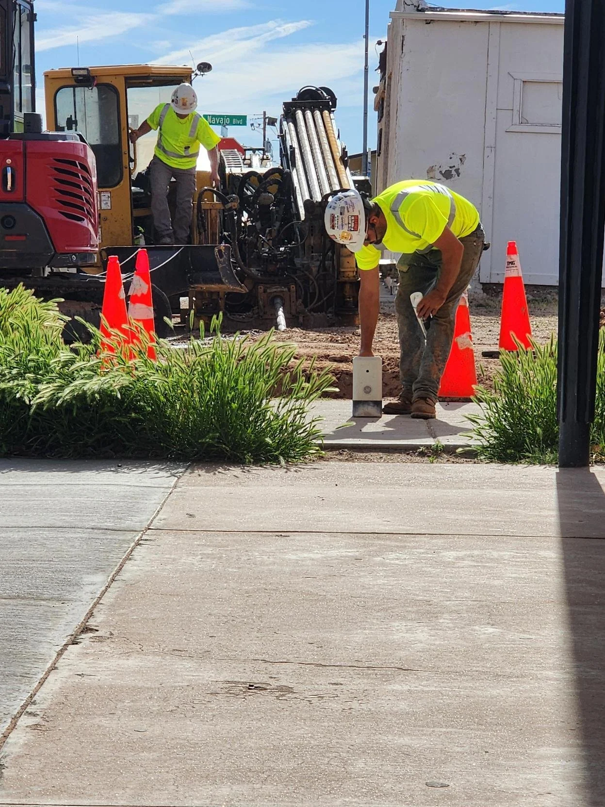 Construction workers are working on a sidewalk, with one operating heavy machinery and another measuring or marking the ground, surrounded by orange safety cones and greenery.