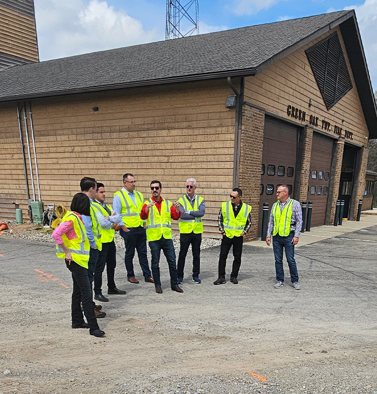 Group of people in yellow safety vests having a discussion outside a wooden building with garage doors.