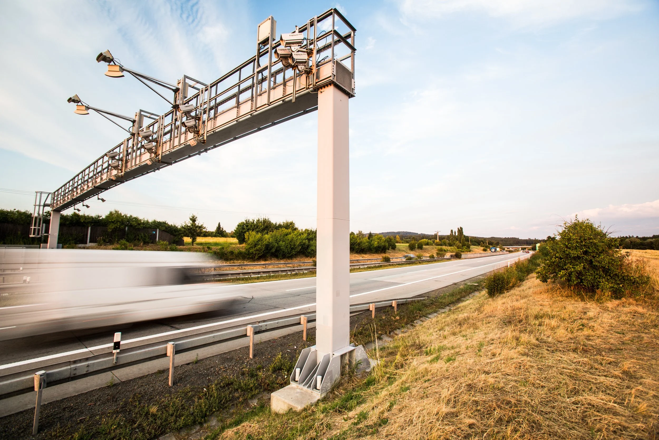 A highway with a highway overpass structure and light sensors, with a blurred white car passing by below, and trees and a partly cloudy sky in the background.