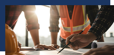 Two construction workers review blueprints on a construction site, with one pointing at the plans, sunlight in background.