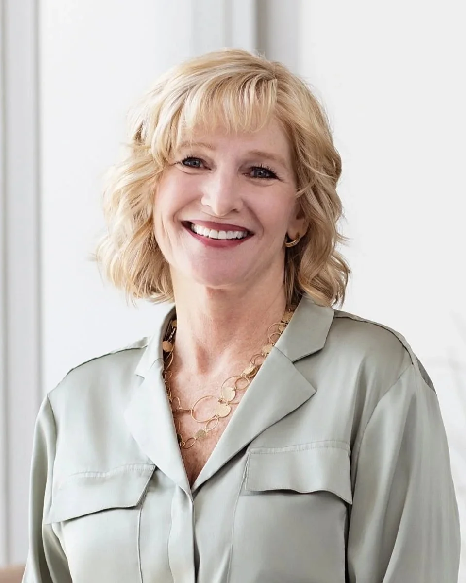 A smiling woman with blonde hair wearing a light-colored blouse and a gold necklace.