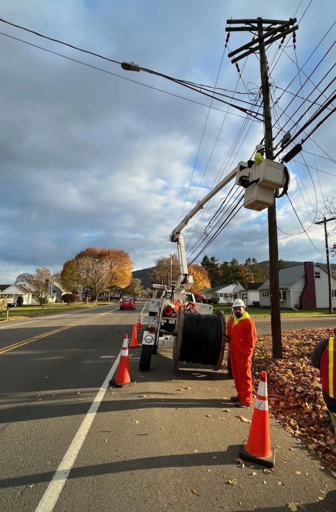 Workers in orange safety gear repairing power lines, with one worker in a bucket lift and another standing on the ground next to a utility pole, traffic cones placed around the work area on a residential street.