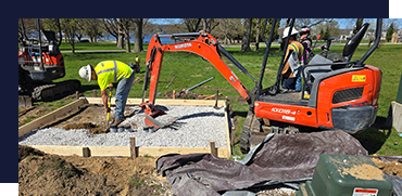 Workers using a compact orange excavator to pour concrete onto a foundation at a construction site.