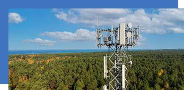 A tall cell tower with antennas stands above a forested area under a partly cloudy sky.