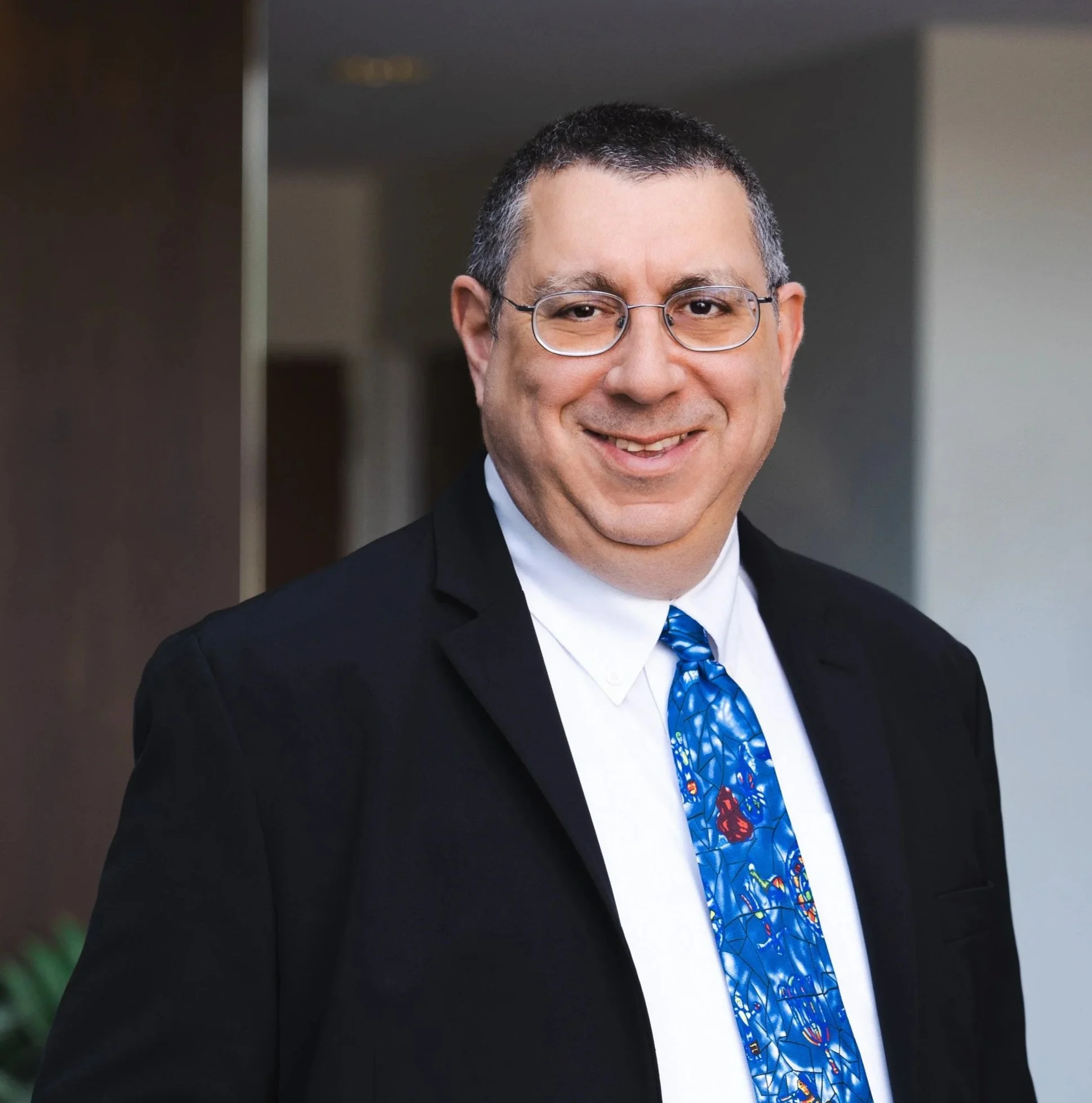 Close-up of a smiling man with short dark hair, glasses, wearing a black suit, white shirt, and a blue tie with a colorful pattern, standing indoors.