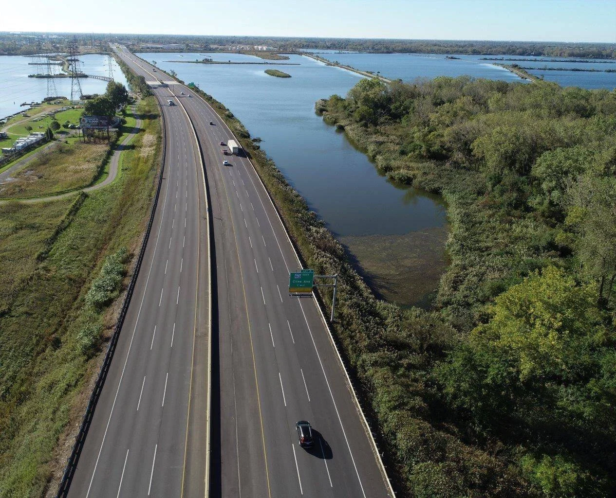 An aerial view of a highway running parallel to a river with dense trees on the right and some buildings and power lines on the left, under a clear sky.