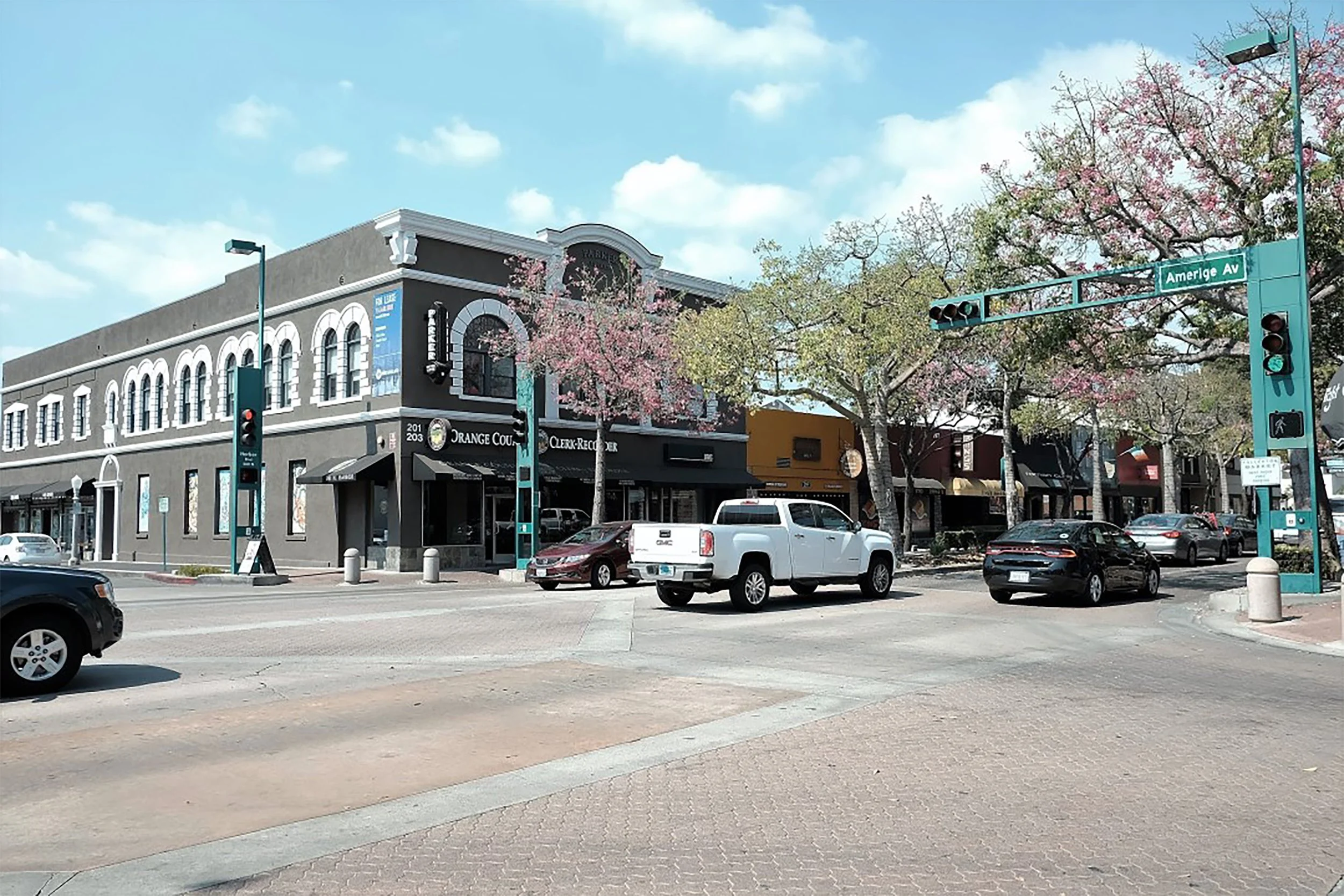 Street view featuring a corner building with a sign for Orange County Clerk-Recorder and a crosswalk. There are several cars parked and a few driving, with a traffic light and street sign for America Ave. The scene has trees with pink blossoms.