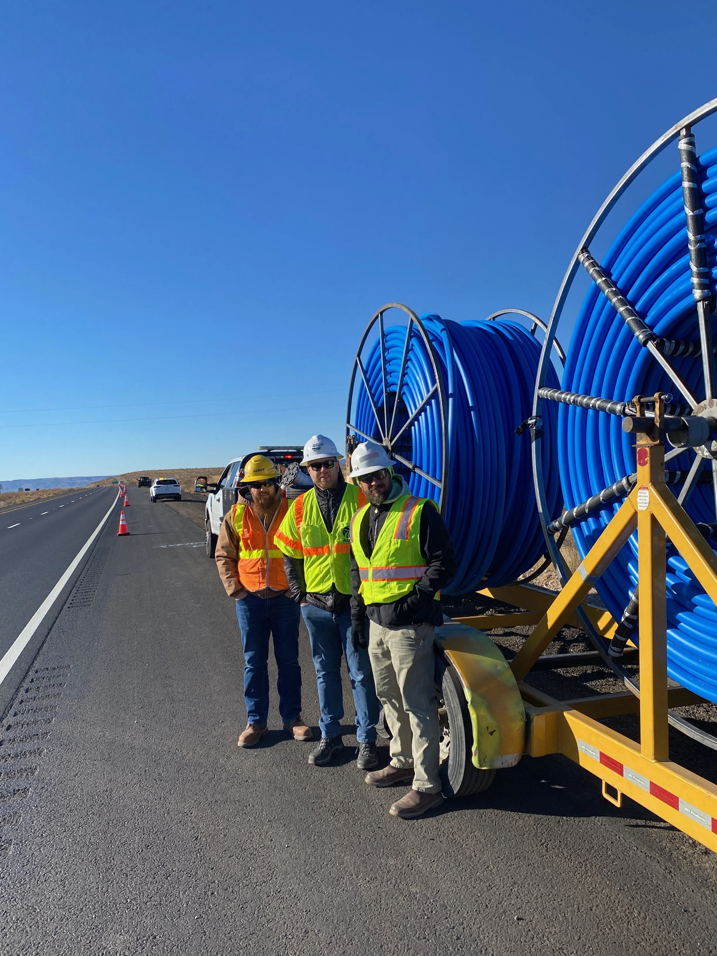 Three workers wearing safety vests and helmets standing beside a large reel of blue pipe on a trailer next to a road under a clear blue sky.