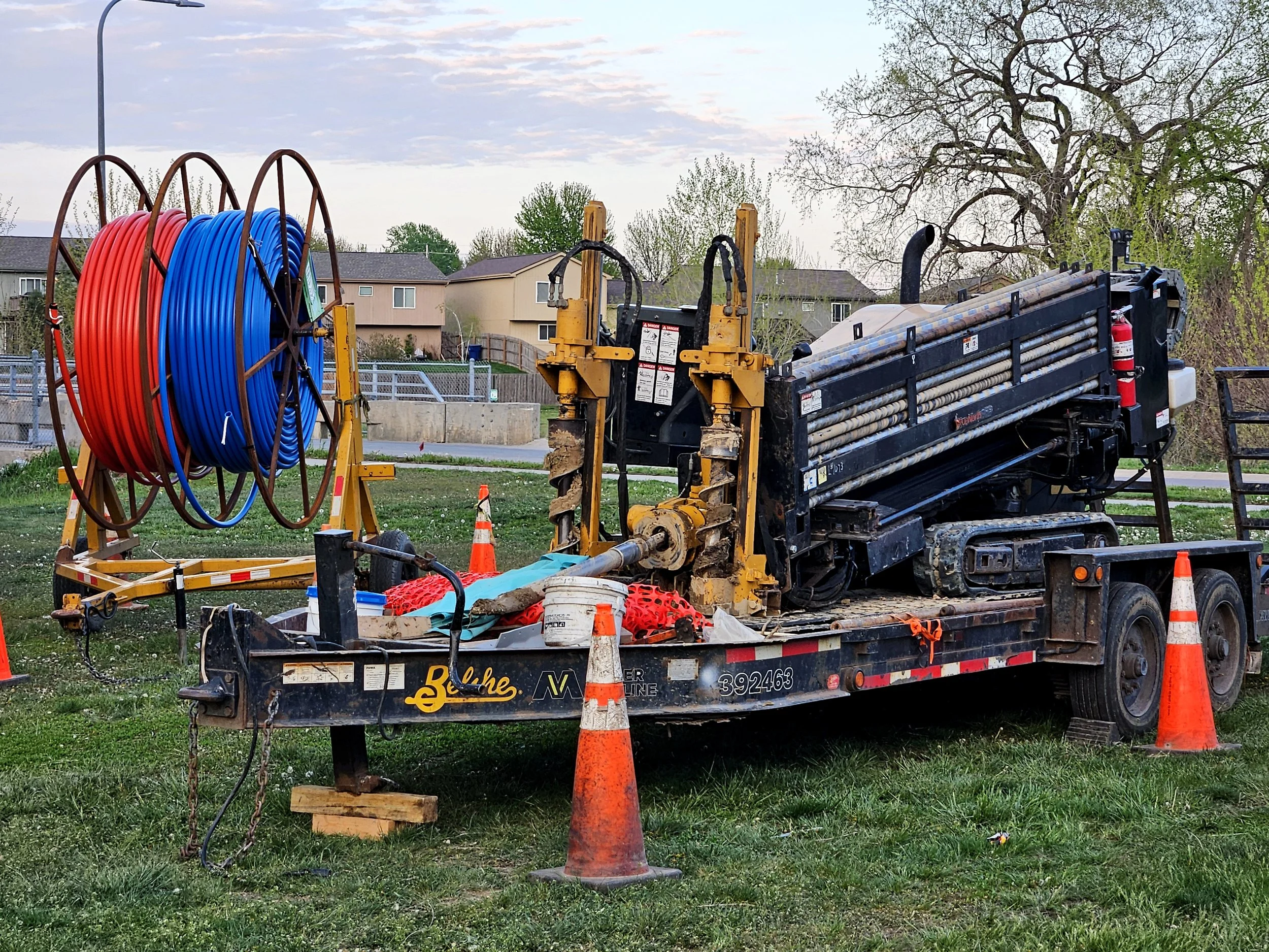 Construction site with a truck, orange cones, and large coiled red and blue hoses on a grassy area. Houses and trees are in the background under a cloudy sky.