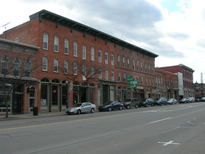 Street view of historic red brick buildings with storefronts and parked cars along the sidewalk under a partly cloudy sky.