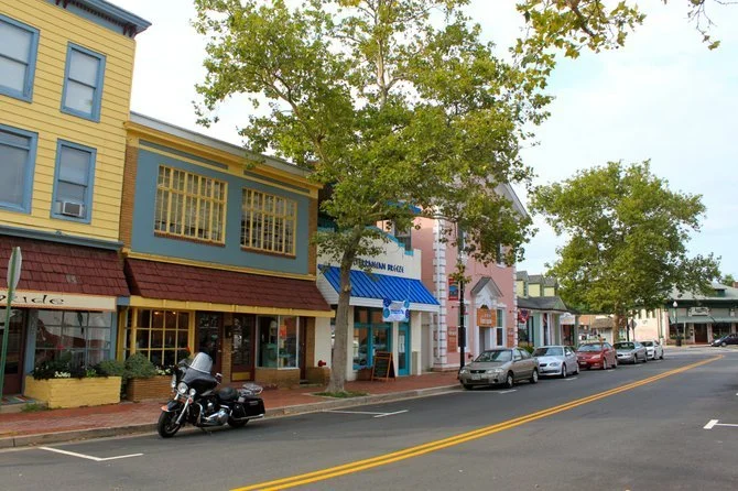 Street view of small-town downtown with colorful buildings, parked motorcycles and cars, sidewalk with trees, and a clear sky.