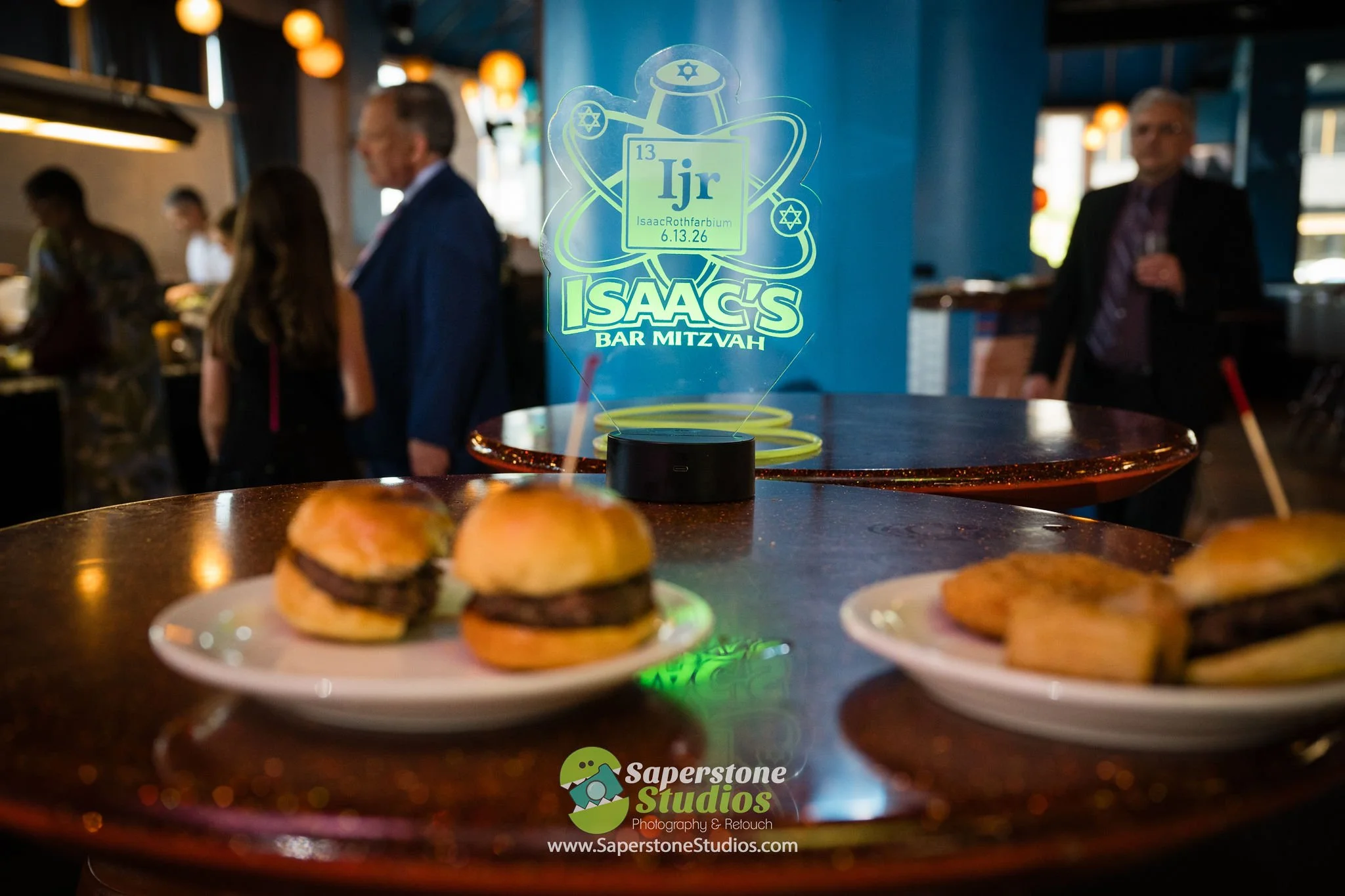 A round table with plates of small sliders and appetizers, with a decorative sign in the background reading 'Isaac's Bar Mitzvah' and a neon menorah, in a dimly lit venue with people in formal attire.
