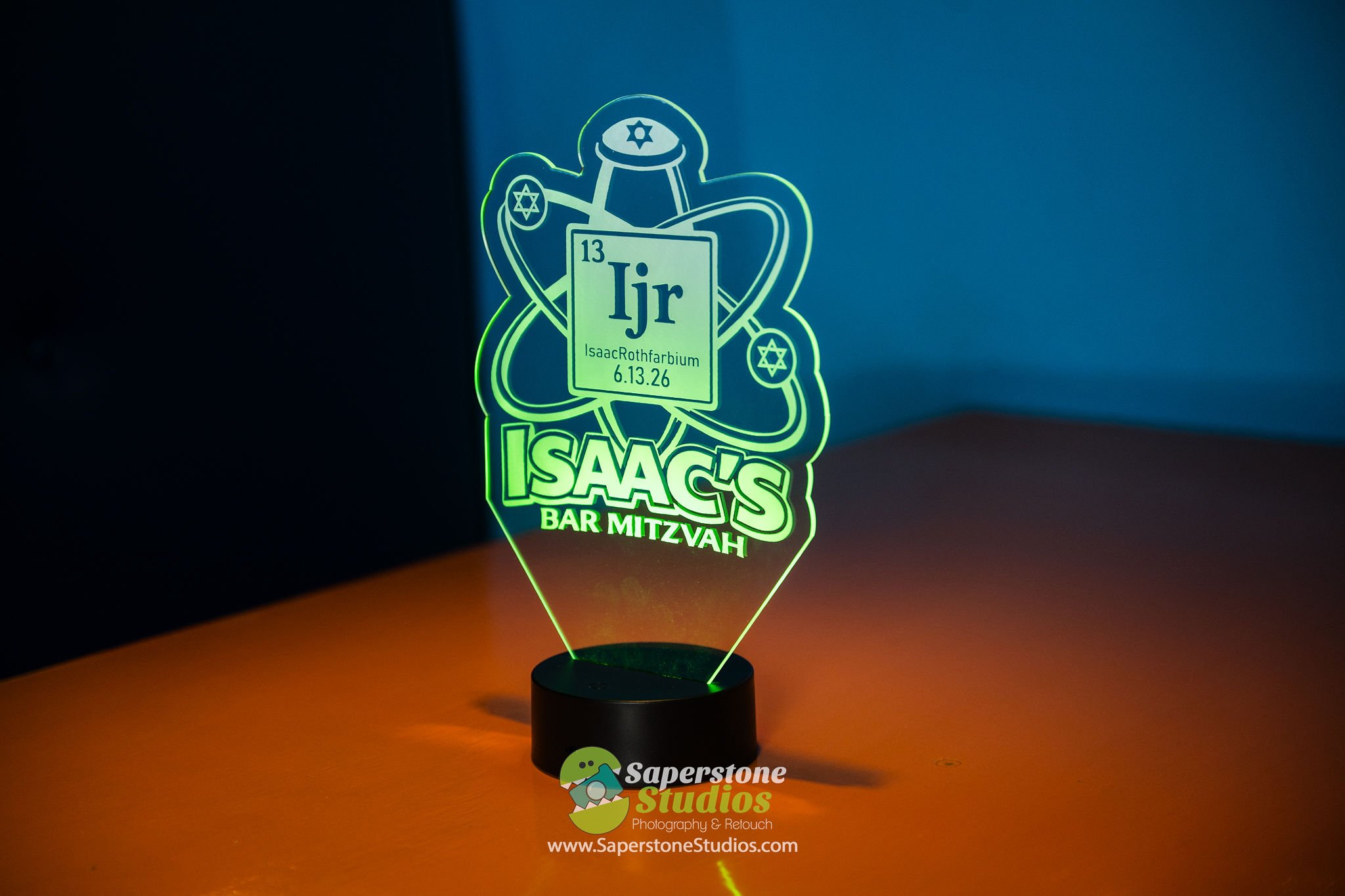 A decorative illuminated acrylic menorah with symbols of Jewish faith, including a Star of David on the top and sides. The menorah features text reading '13 Jr Isaac Rothfarbium 6.13.26' and a logo for 'Isaacs Bar Mitzvah' at the bottom. The menorah 