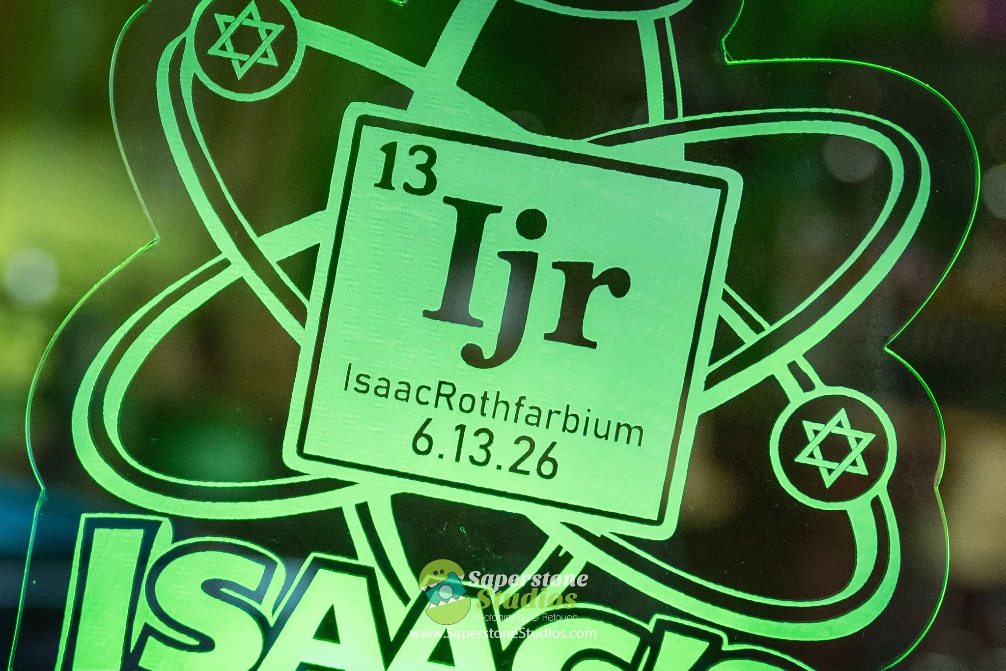 A glowing green neon sign featuring a design with various elements including a six-pointed star, a stylized teacup, and text displaying an event date of June 13 and 26, along with the location Isaac Rothfarbium. The sign has a vibrant, luminous appea