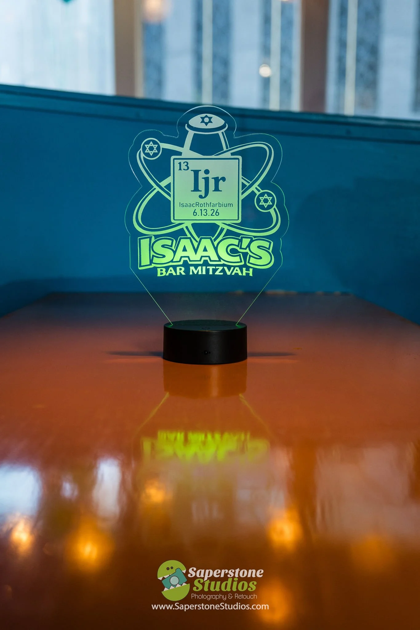 A decorative LED light featuring the periodic table element iodine (I) with the symbol Ijr, the date 6.13.26, and the name Isaac Rothfarbium. The design includes atomic orbitals and star symbols, with the text 'Isaac's Bar Mitzvah.' The logo of Saper