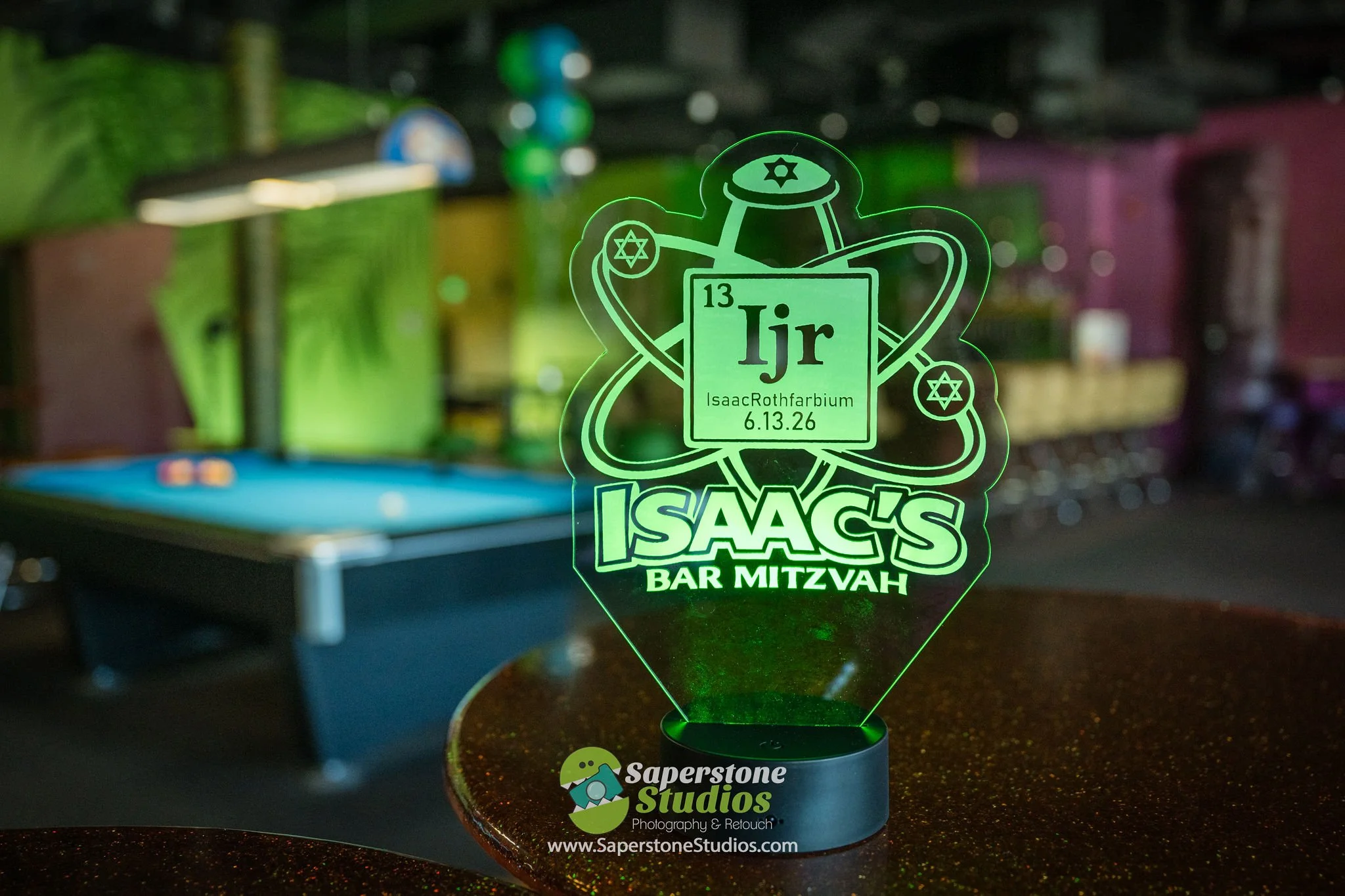 Neon sign with the periodic table symbol for iridium (Ir), displaying the date 6.13.26, referencing Isaac Rothfarbium, at a bar mitzvah celebration, with a billiards table and colorful background in a dimly lit room.