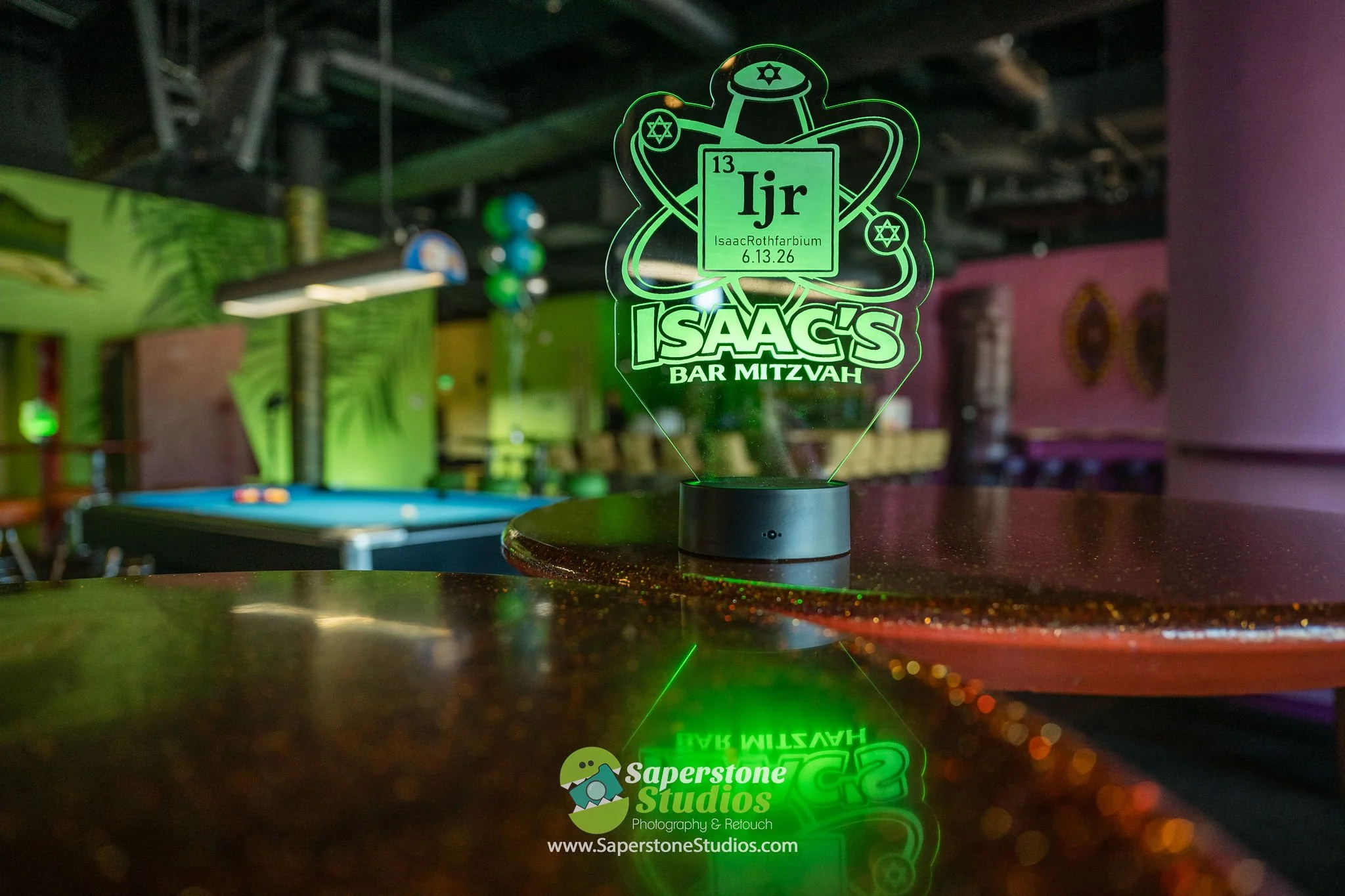 A decorative green acrylic sign with scientific elements, featuring the name 'Isaac Rothfarbium', the date 6.13.26, and the initials 'Ijr', placed on a dark table in a colorful lounge or bar area.