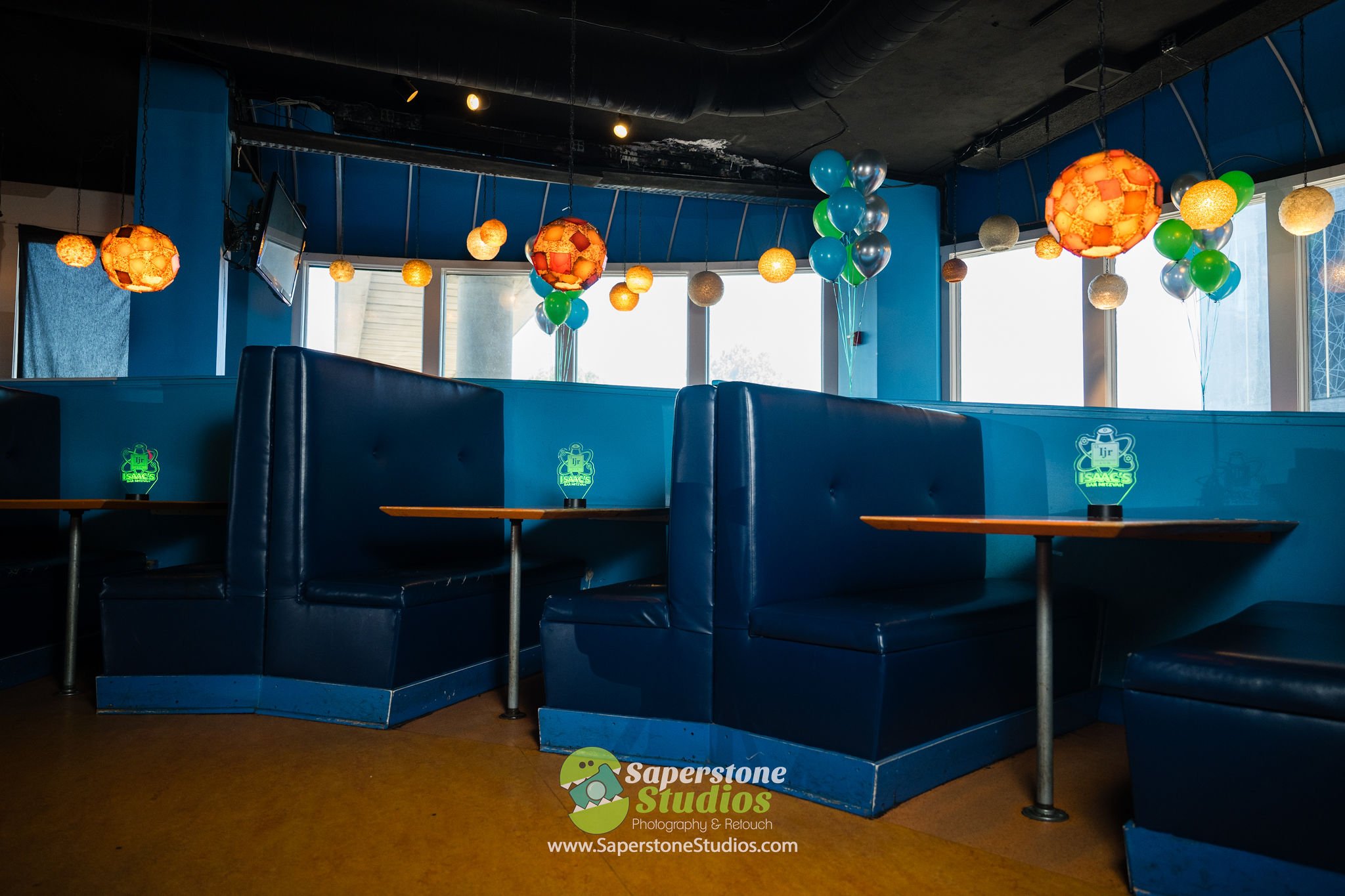 Inside a restaurant with blue booths, decorative hanging lamps, and clusters of balloons near windows, with a television mounted on the wall.