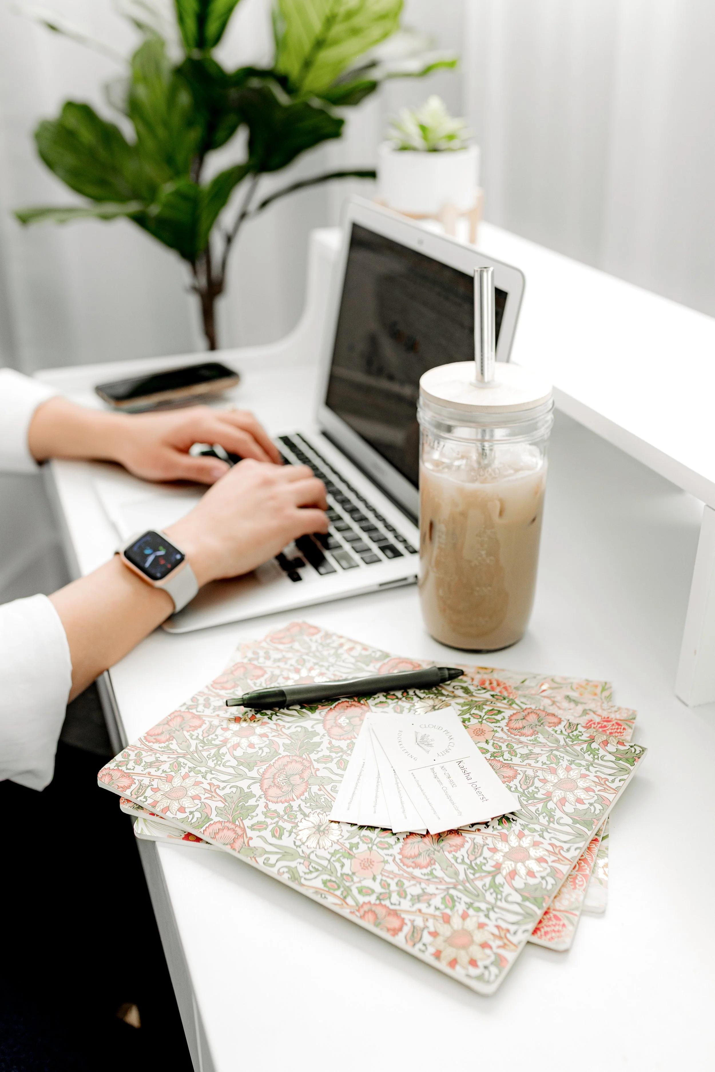 Kaisha Jokerst working on a laptop with a smartwatch on her wrist, an iced coffee in a jar with a straw, floral-patterned notebooks, a pen, and some business cards on a white desk. A potted plant is in the background.