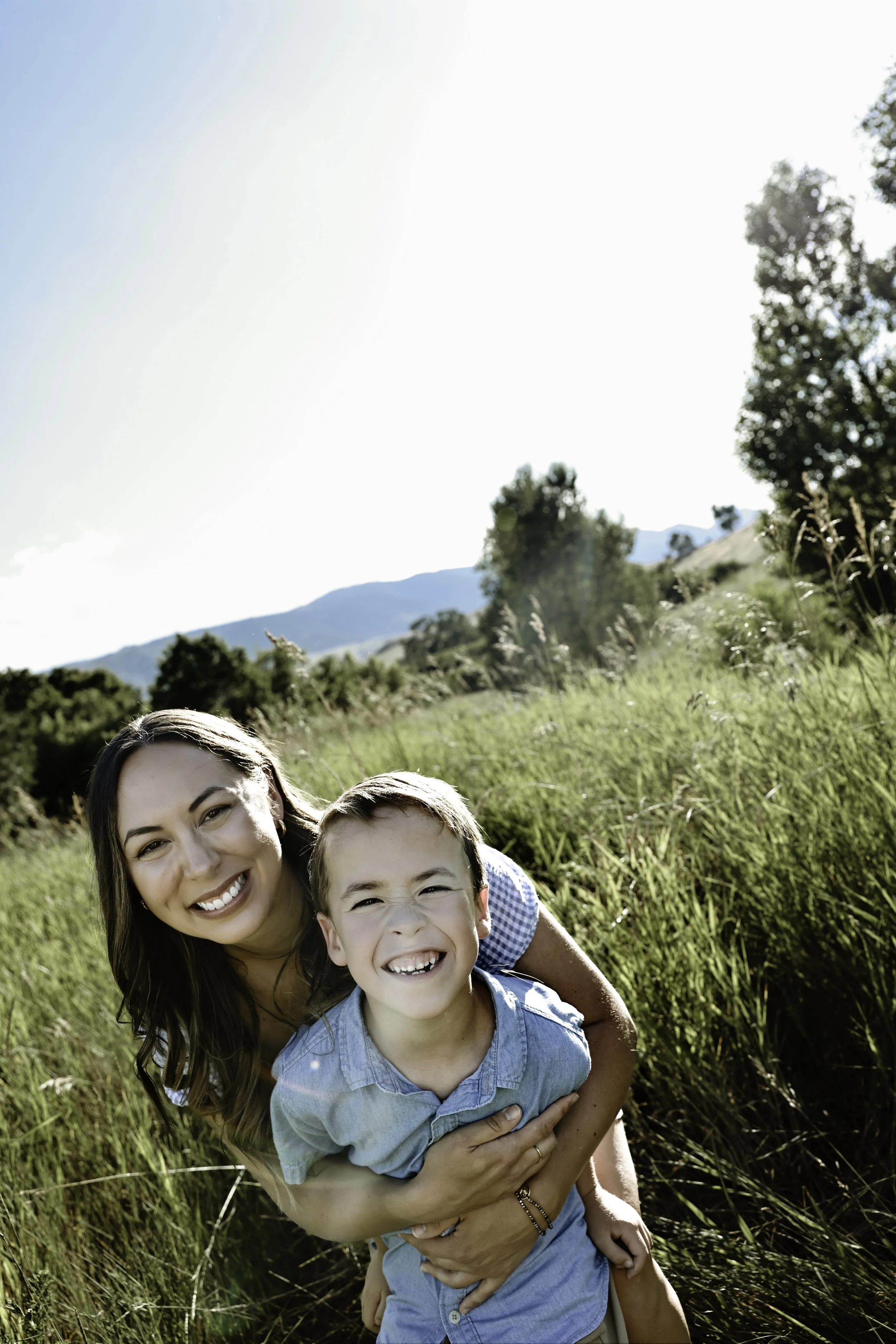 Kaisha Jokerst and son with bright smiles, hugging each other outdoors in a grassy field with trees and mountains in the background, under a clear, sunny sky.