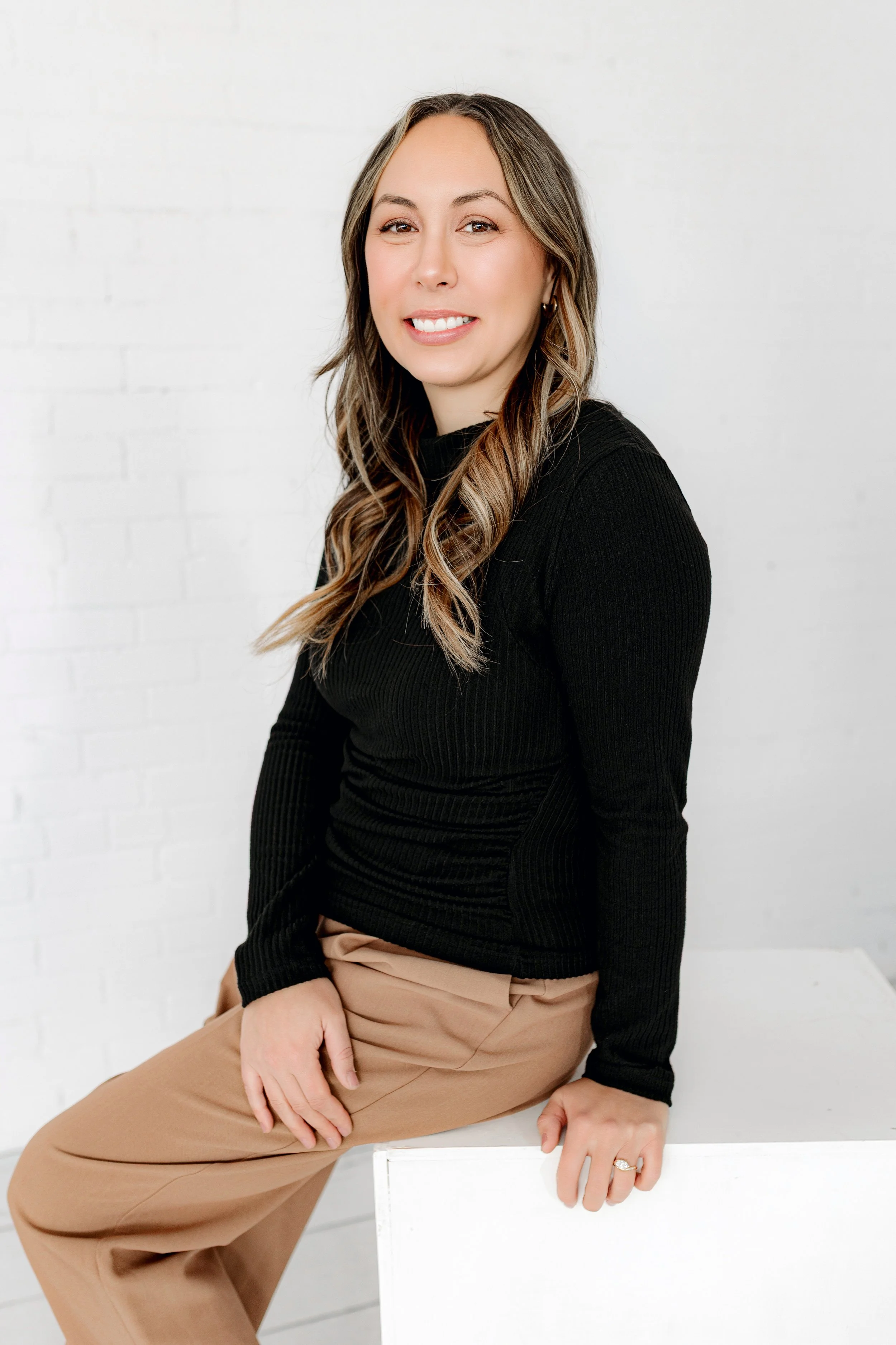 Portrait of Kaisha Jokerst, sitting on a white surface against a white brick wall, smiling at the camera.