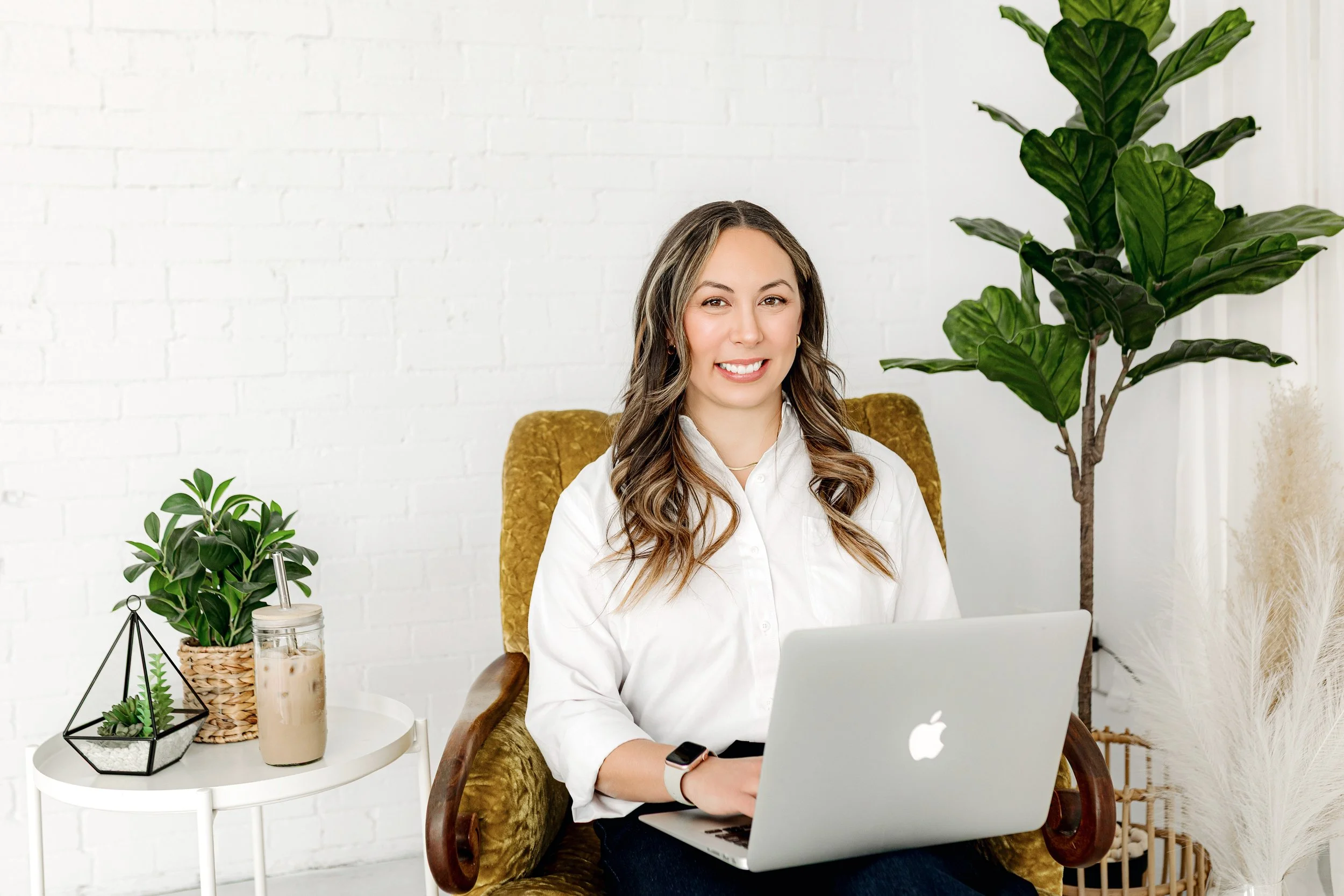 Kaisha Jokerst sitting on a vintage mustard-colored armchair using a MacBook, with plants and decorative items on a small white side table and a large plant behind her, in a bright room with white brick wall.