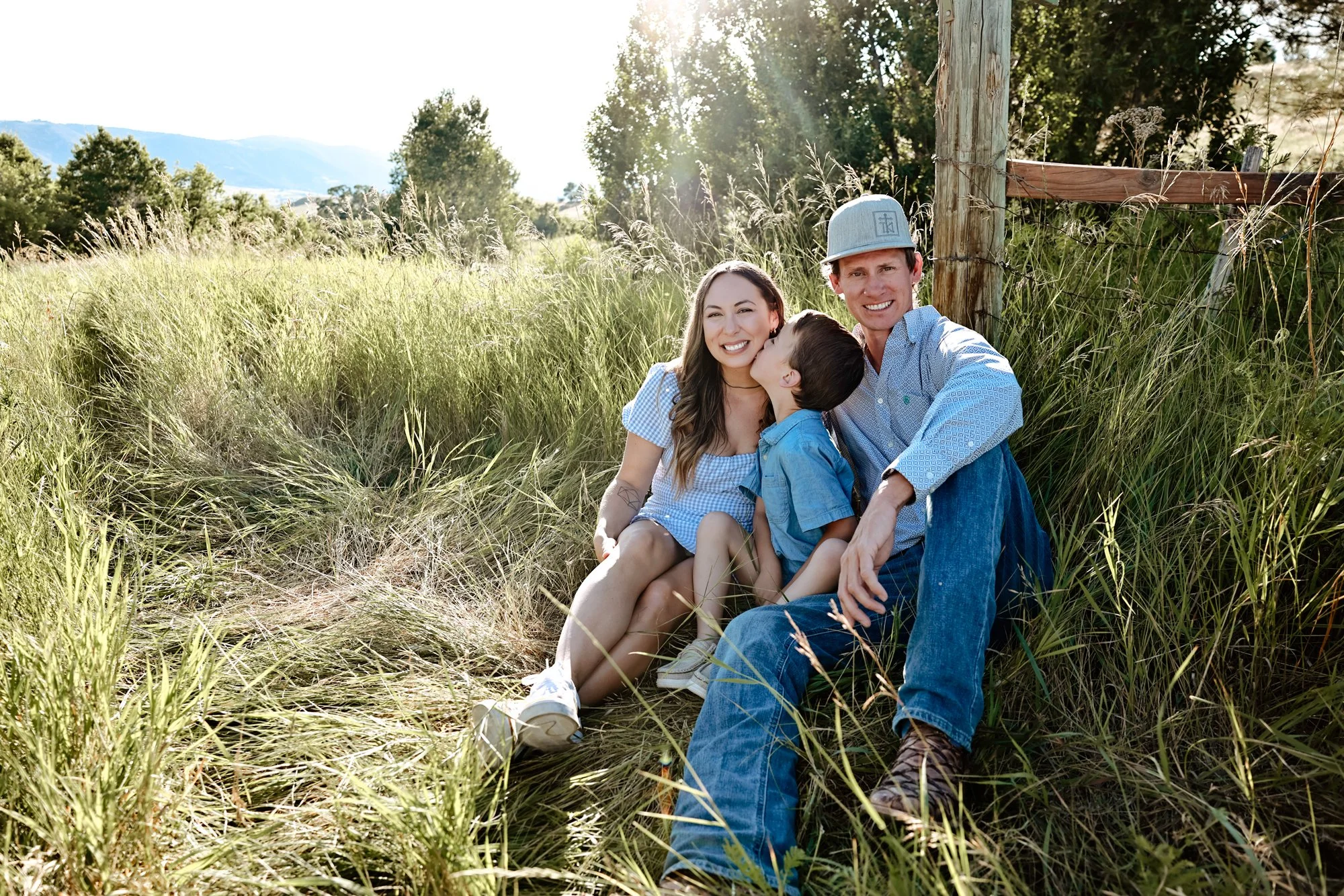 A family of three sitting in tall grass outdoors, smiling and enjoying the sunny day. The woman is on the left, with long brown hair, wearing a light blue dress. The boy is in the middle, giving the woman a kiss, wearing a blue shirt. The man is on the right, wearing a gray cap, a light blue shirt, and jeans.