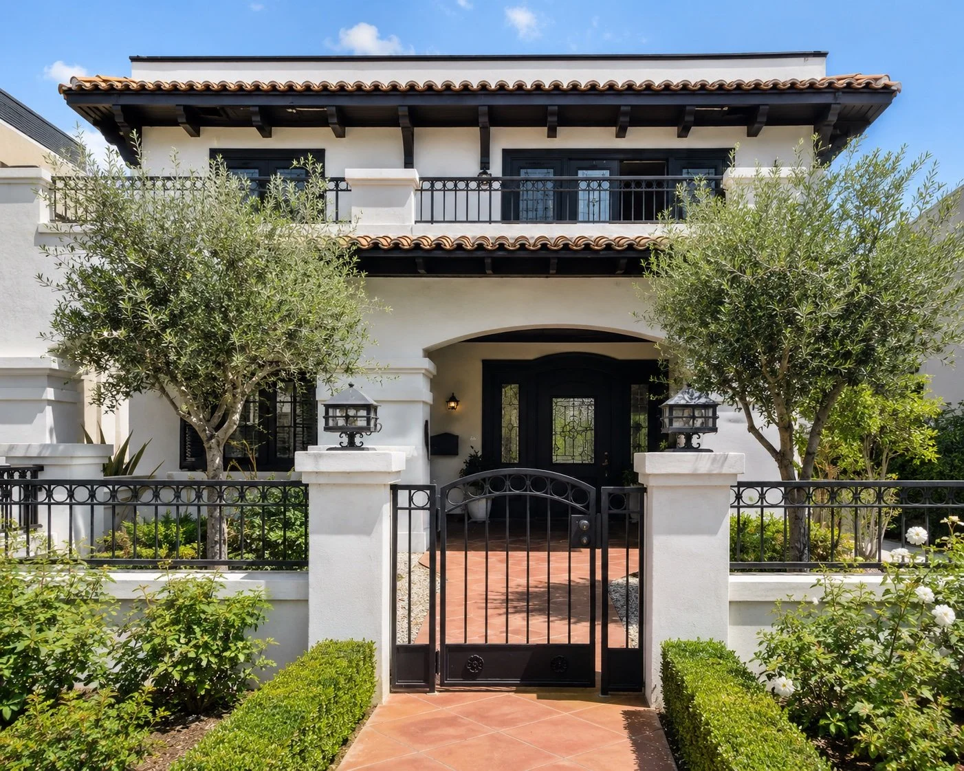 A two-story house with white walls, black accents, and a tiled roof. There is a front garden with trimmed bushes, trees, a black metal gate, and stone lantern-style lights near the entrance. The sky is blue with a few clouds.