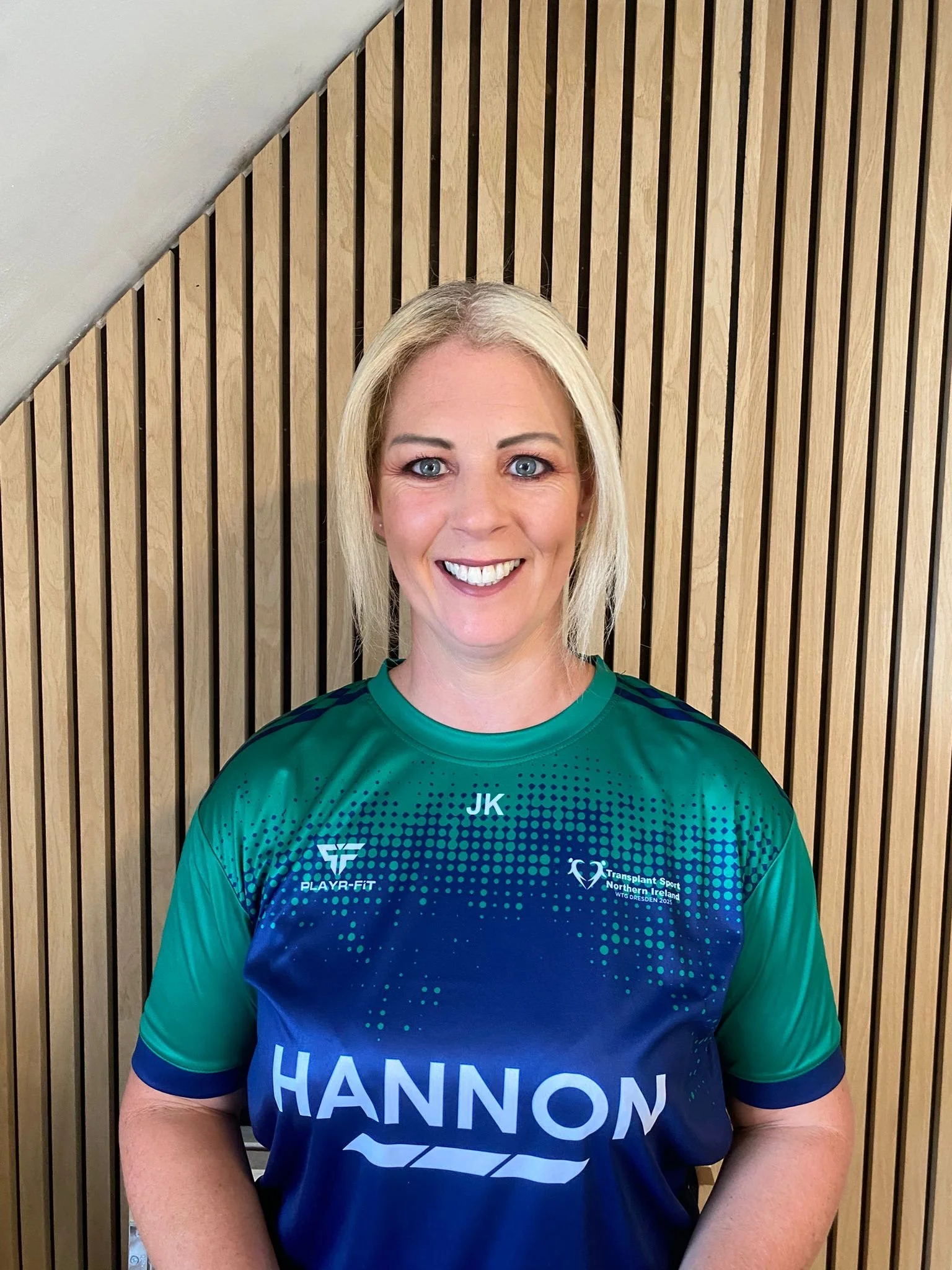 A woman with blonde hair and blue eyes wearing a blue sports jersey with the name 'HANNON' and logos, standing in front of a wooden slatted wall.