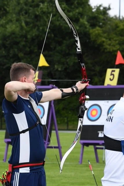 A man practicing archery outdoors, aiming with a bow and arrow at a target.