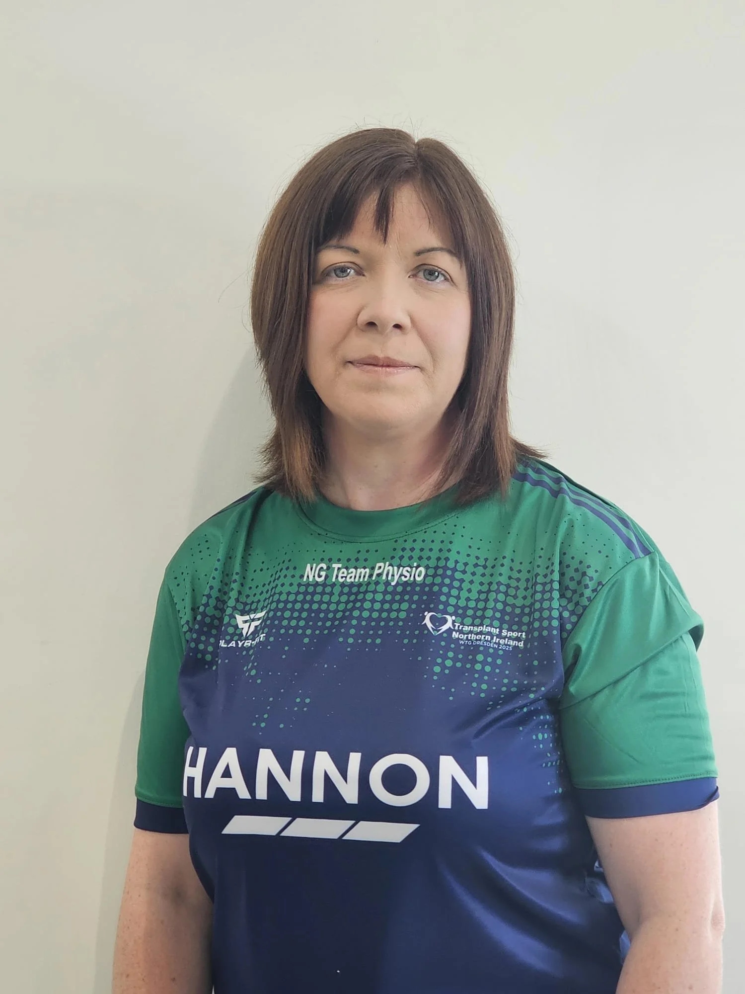 A woman with shoulder-length brown hair and blue eyes wearing a green sports shirt with logos and the name 'Hannon' on it, standing against a plain light-colored wall.