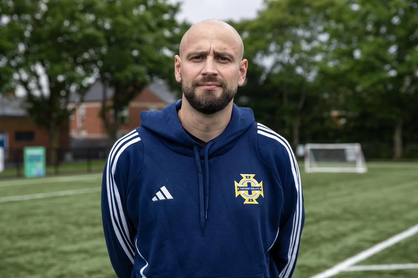 A man with a beard and shaved head wearing a blue Northern Ireland football hoodie standing on a football field with trees and goalposts in the background.