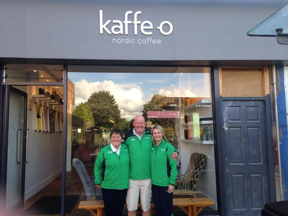Three smiling people wearing green jackets standing in front of a coffee shop named Kaffe o, Nordic Coffee, outside a building with trees and clouds reflected in the glass window behind them.