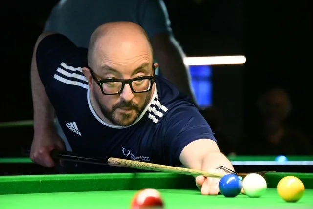 Man with glasses playing billiards, aiming to strike the cue ball.