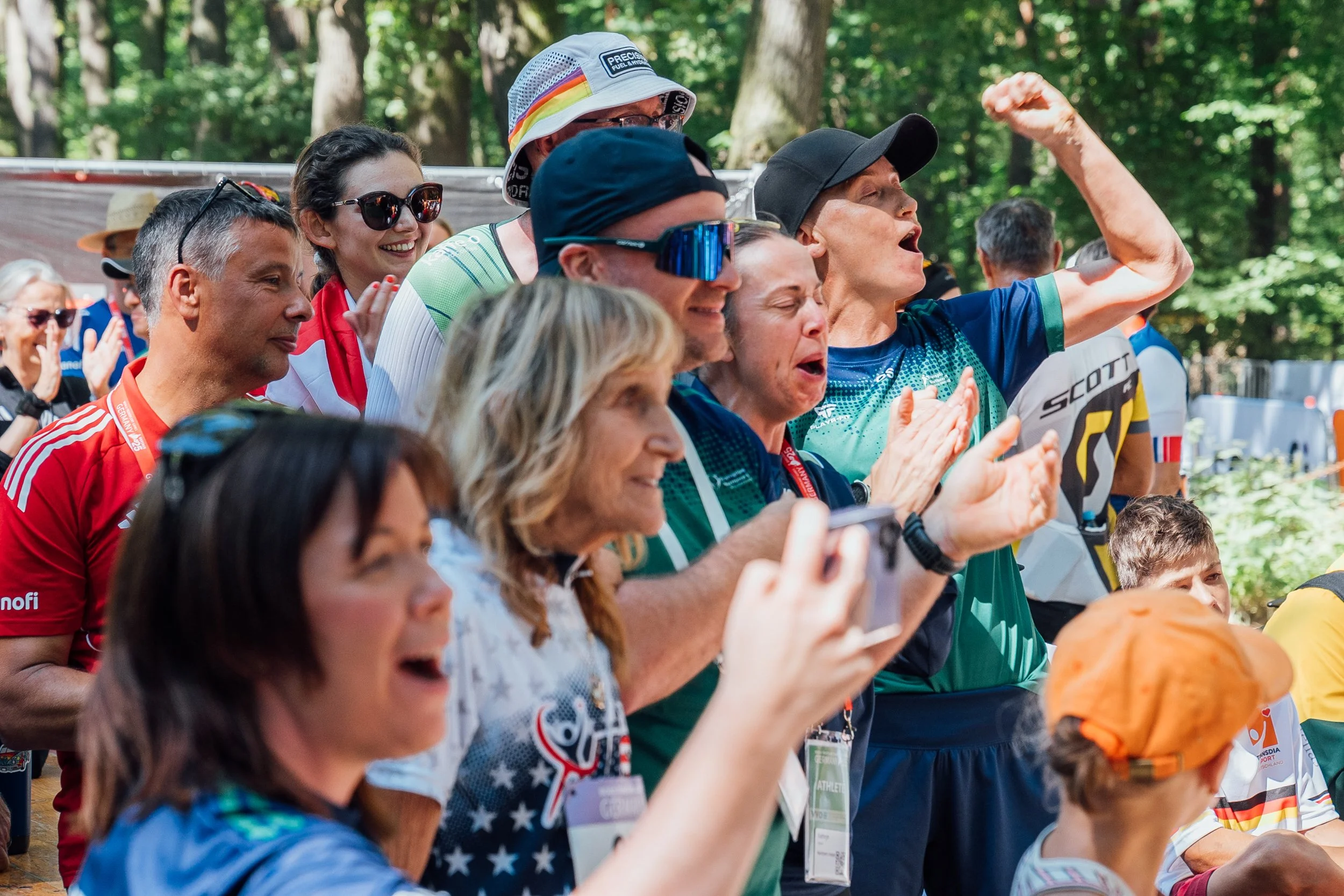 A group of people outdoors, celebrating, some cheering and smiling, some wearing sports gear and sunglasses.