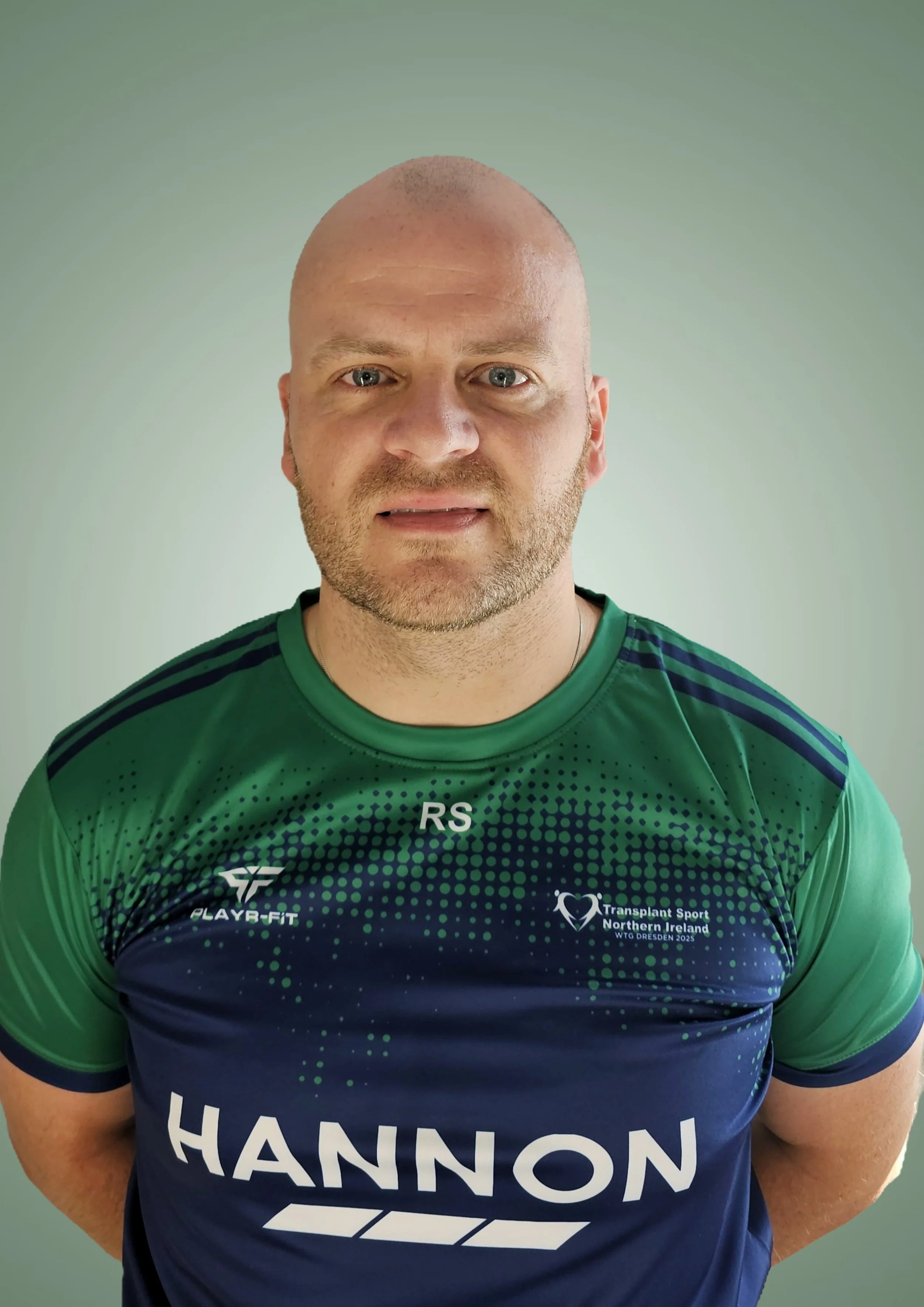 A man with a shaved head and beard wearing a green and blue sports jersey with sponsor logos and the word 'HANNON' printed on it.