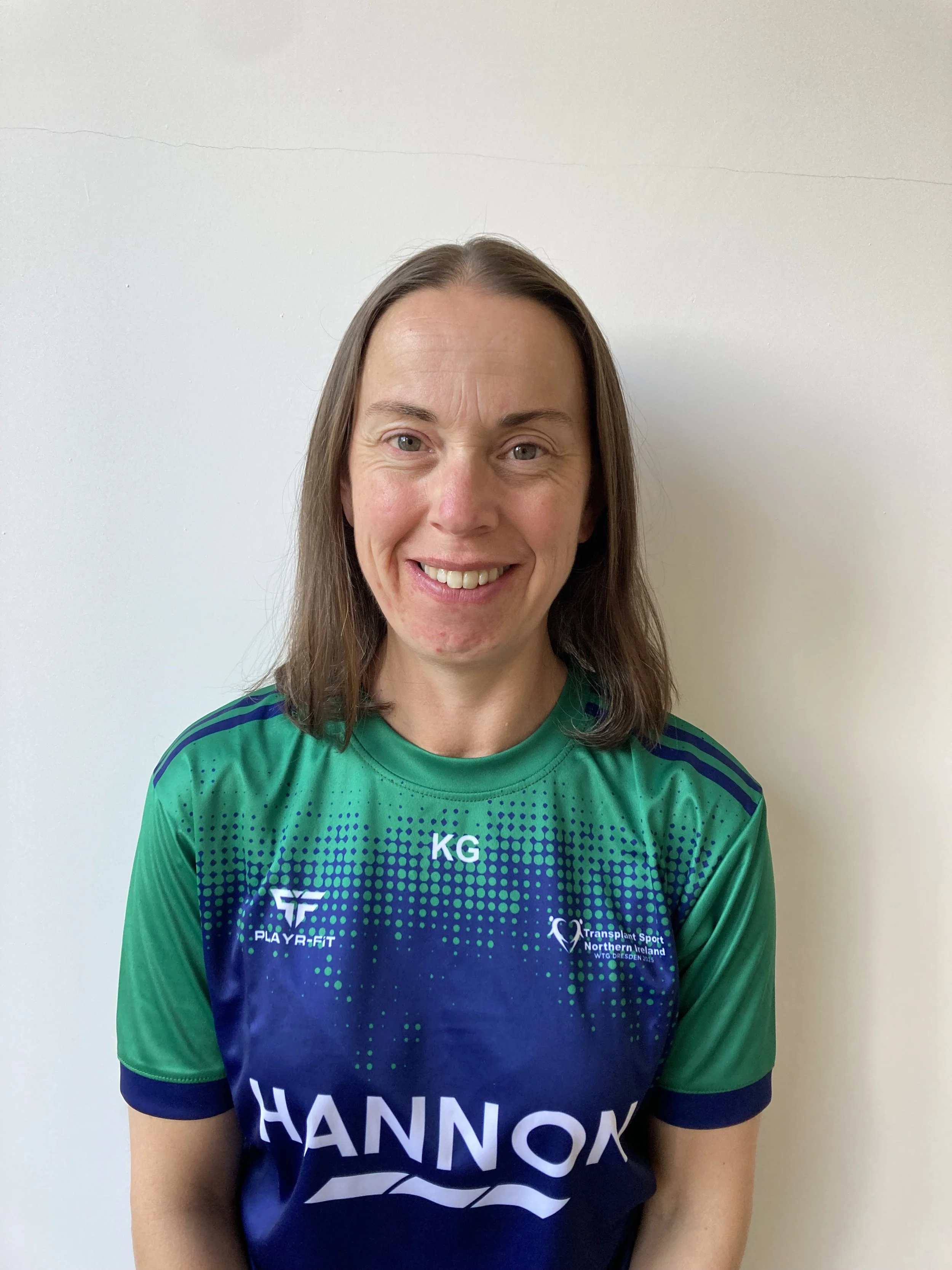 A woman with long brown hair wearing a blue and green sports shirt standing against a plain white wall, smiling at the camera.