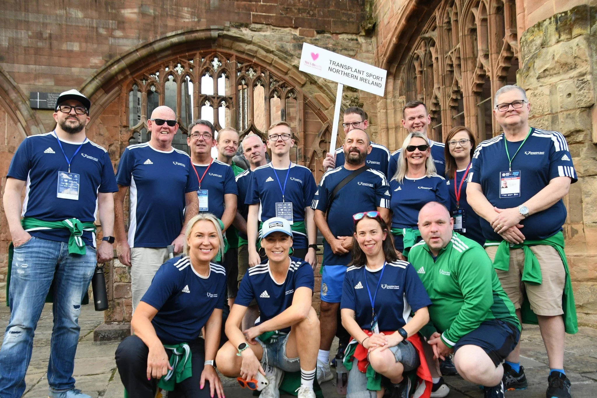 Group of people wearing sports shirts with medals, some holding a sign that reads "Transplant Sport Northern Ireland," standing in front of a historic building with arched windows and stone walls.