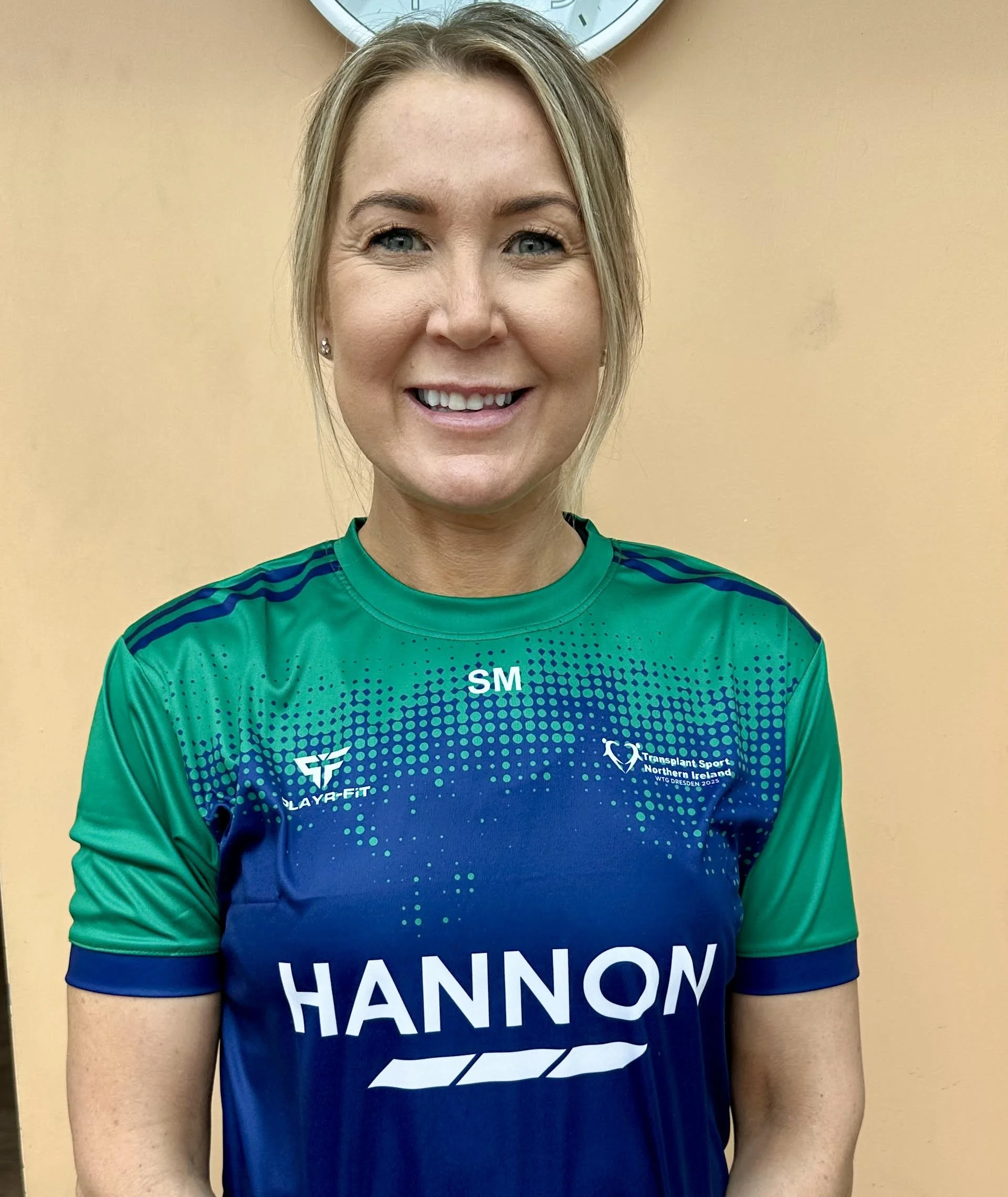 A woman in a sports jersey with the name 'Hannons' and the logo 'Transplant Sport Northern Ireland' standing against a peach-colored wall, with a clock above her.