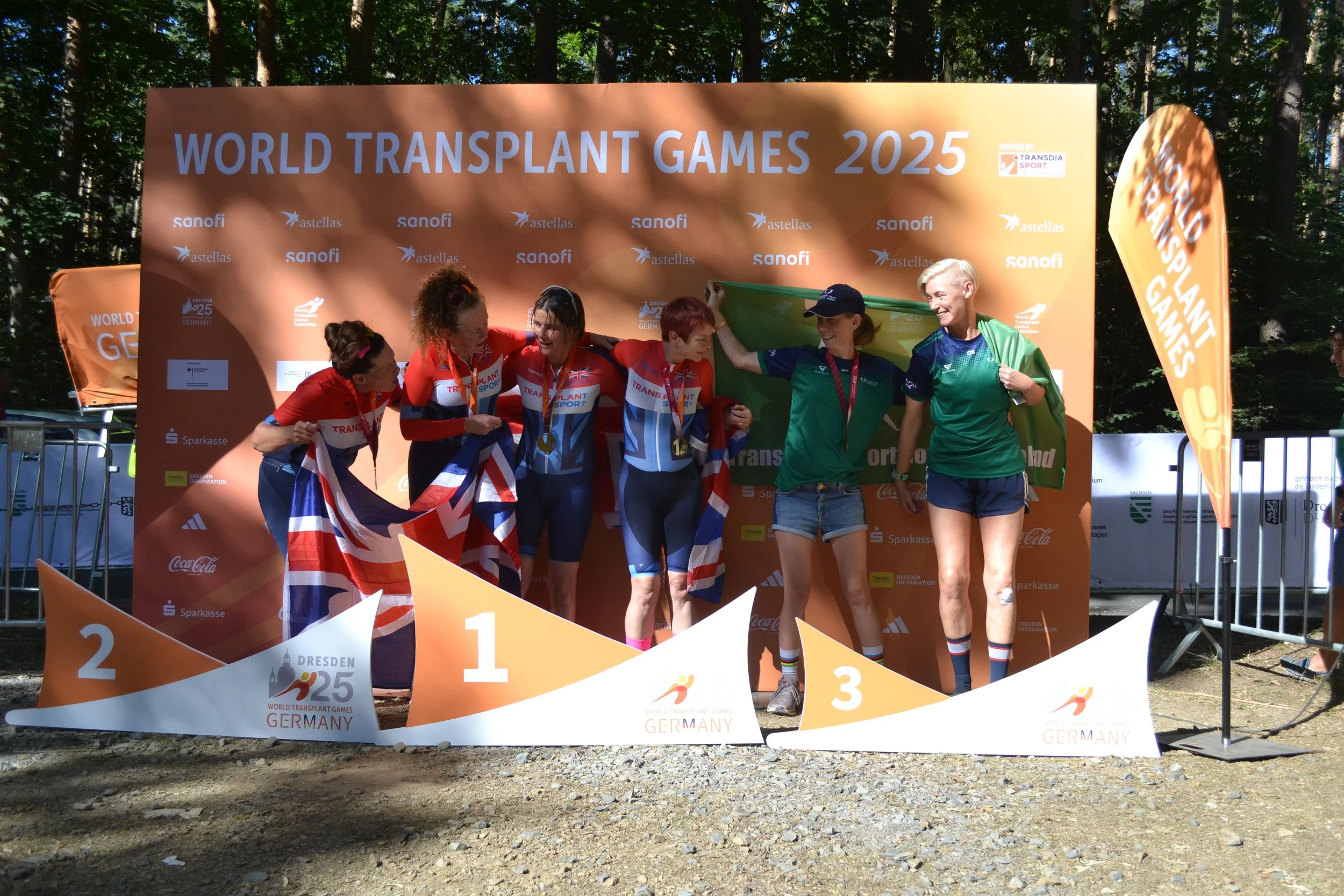 Athletes on a winner's podium at the World Transplant Games 2025 in Germany, surrounded by sponsorship banners and trees in the background, celebrating their medals and achievements.