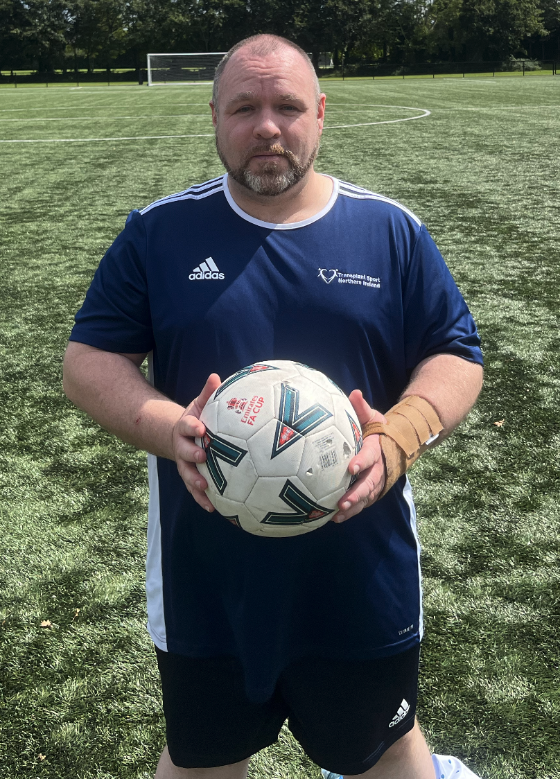 A man standing on a soccer field holding a soccer ball, wearing a blue sports jersey with a logo on the chest and Adidas stripes on the shoulders, with a soccer goal in the background.