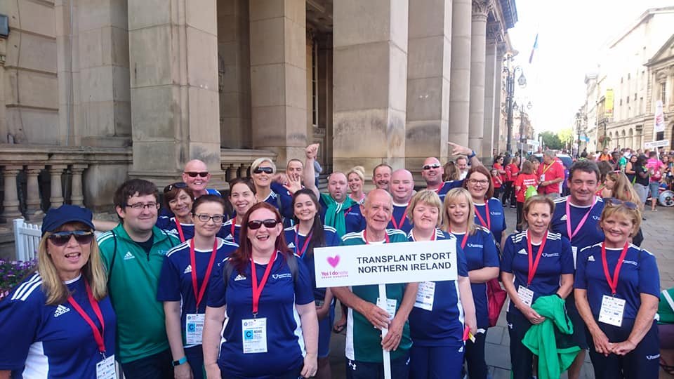 A group of people wearing blue and green sports shirts with medals, gathered outdoors in front of a historic building, some holding a sign that reads 'Transplant Sport Northern Ireland.'