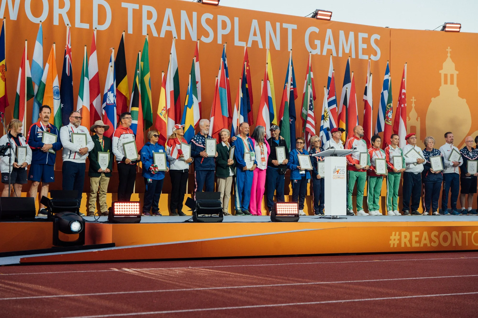Group of people standing on stage during the World Transplant Games awards ceremony, holding framed certificates. The background features multiple flags and a large orange banner with the text 'World Transplant Games.'