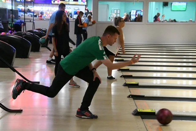 A man in a green shirt is bowling at a bowling alley, preparing to release a purple bowling ball down the lane. Other people are visible in the background, some watching and some in line.