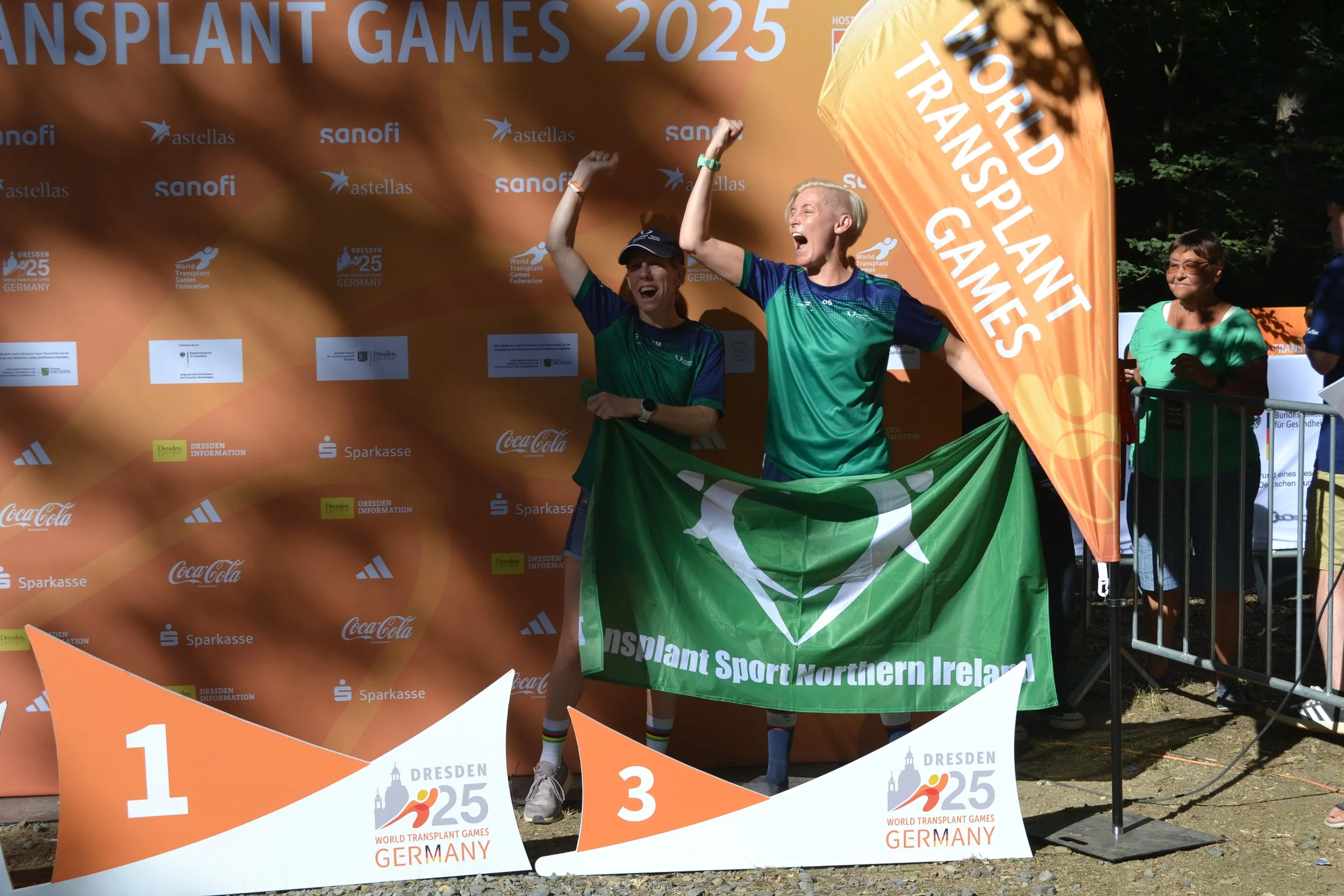 Three athletes standing on a winners' podium celebrating at the World Transplant Games 2025 in Germany. The first-place winner is a woman wearing a green and blue athletic uniform, holding a flag, with her arms raised in victory. The second-place ath