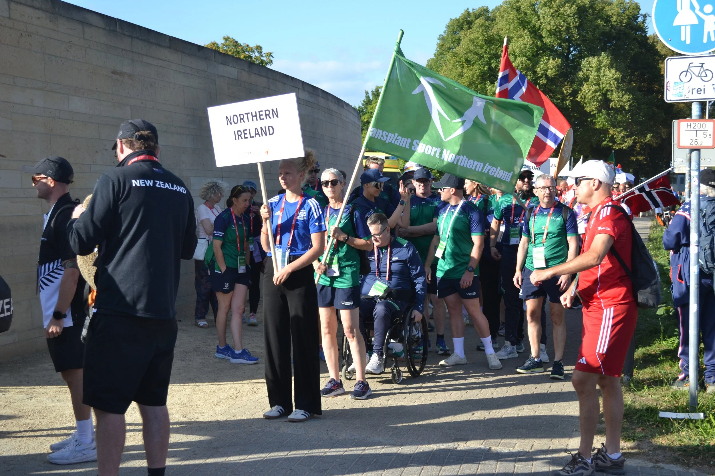 A group of people participating in a parade or event, some wearing sportswear, holding flags from Northern Ireland, New Zealand, and others. The scene is outdoors, with trees and a wall in the background, and various signs visible including a blue pe